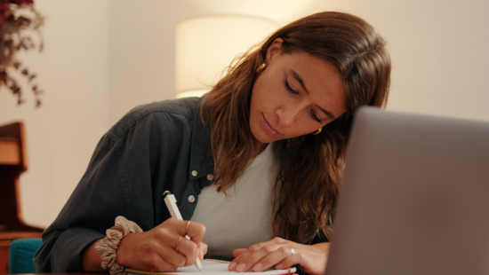 A person takes notes in a notebook next to an open laptop, representing online learning and professional certification.